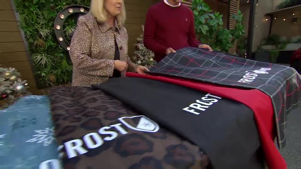 A woman in a leopard-print jacket and a man in a burgundy sweater are displaying a stack of blankets. The blankets, each with "FROST" printed on them, are in various patterns and colors.

