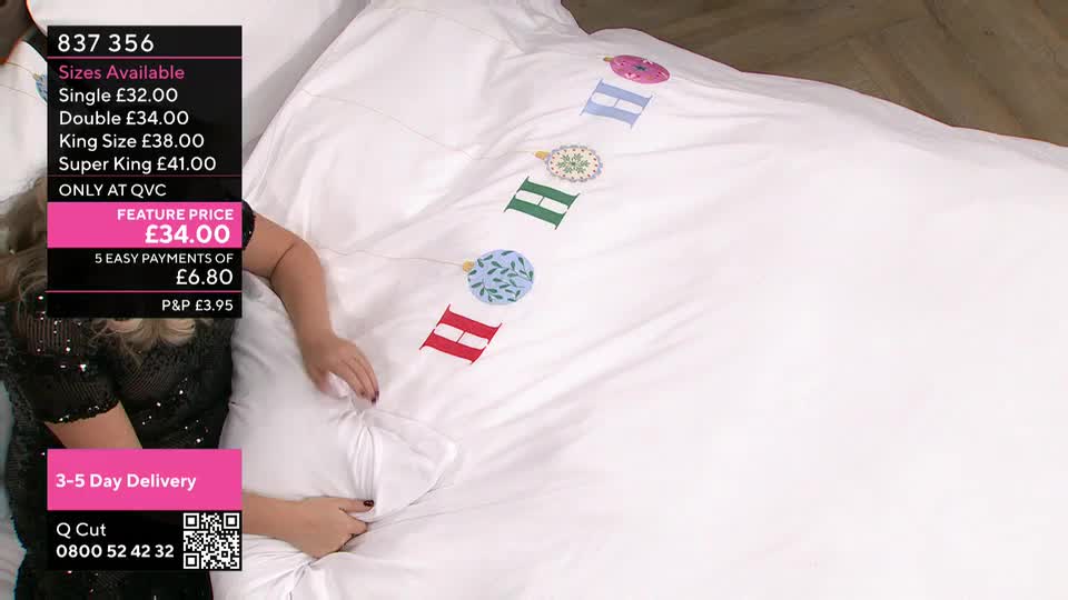 A presenter on QVC Extra is touching a white duvet cover decorated with colorful, stitched ornaments. The price and sizes are displayed on the screen in the United Kingdom.
