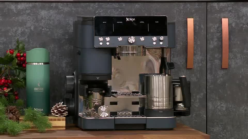 A Ninja coffee machine sits on a wooden surface, ready for demonstration on QVC Extra from the United Kingdom. Beside it, a green travel mug and festive greenery add a touch of color to the scene.
