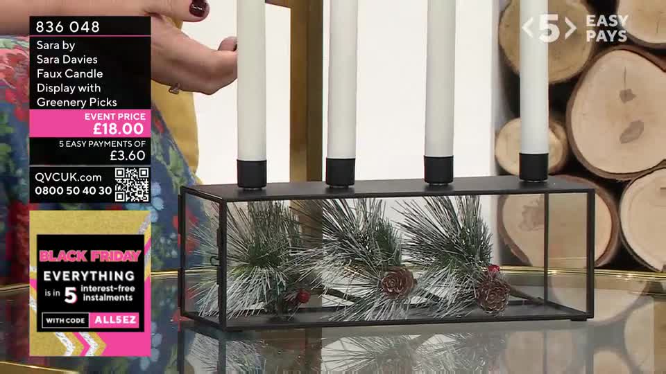 A black metal candle holder with four white faux candles sits on a glass surface. Inside the holder, artificial pine branches and pine cones are arranged.

