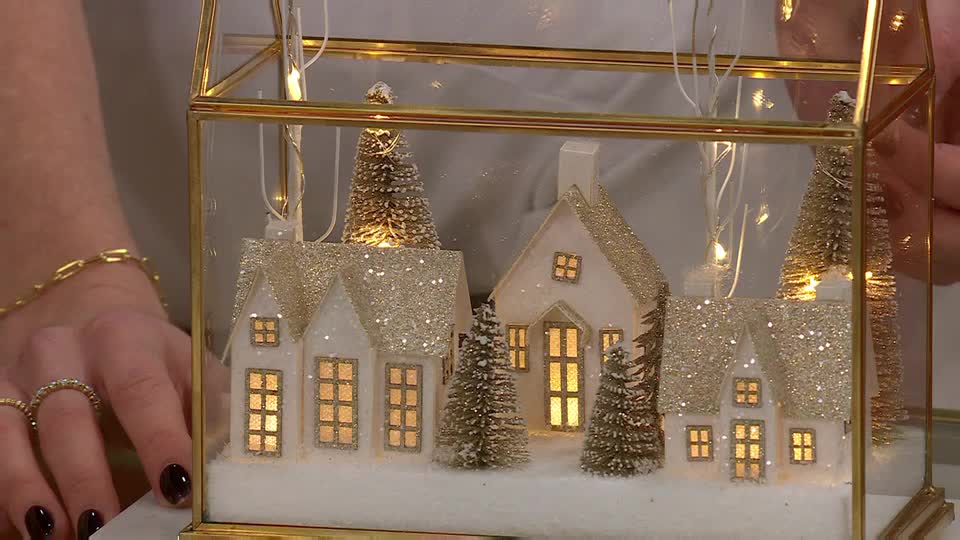 Inside a glass terrarium, tiny white houses with gold glitter roofs and lit windows sit nestled among miniature trees. A presenter, adorned with gold jewelry, gently holds the display, showcasing it for the QVC Extra viewers in the UK.
