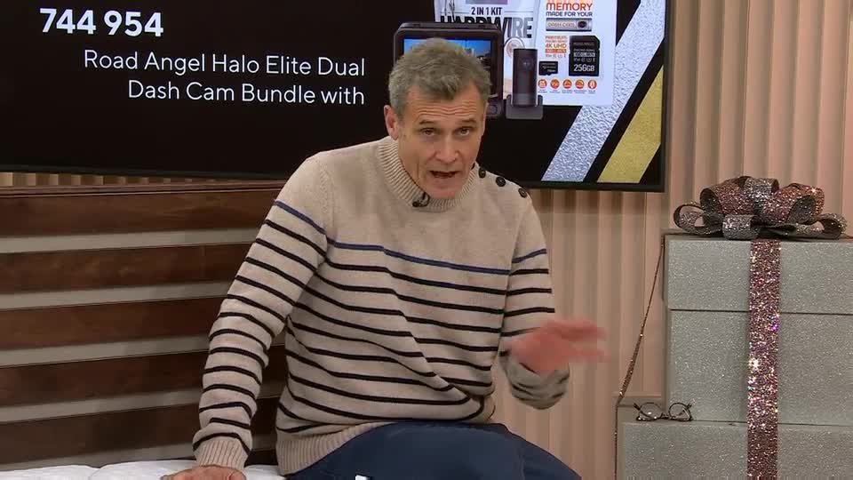 The host on QVC Extra in the United Kingdom gestures with his right hand as he speaks. Behind him, a screen displays the product and its price.
