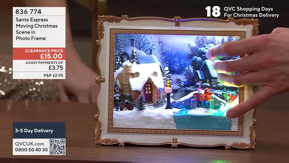 A hand is gesturing toward a miniature winter scene inside a decorative frame. The scene is lit with blue and green lights, and the frame is being presented on QVC Extra in the United Kingdom.
