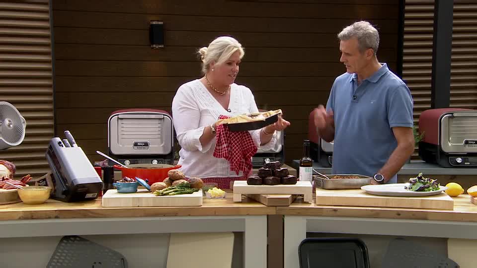 A woman in a white blouse holds up a cooked dish, gesturing towards it with a red-checkered cloth. The man beside her points, seemingly explaining the food to the audience.
