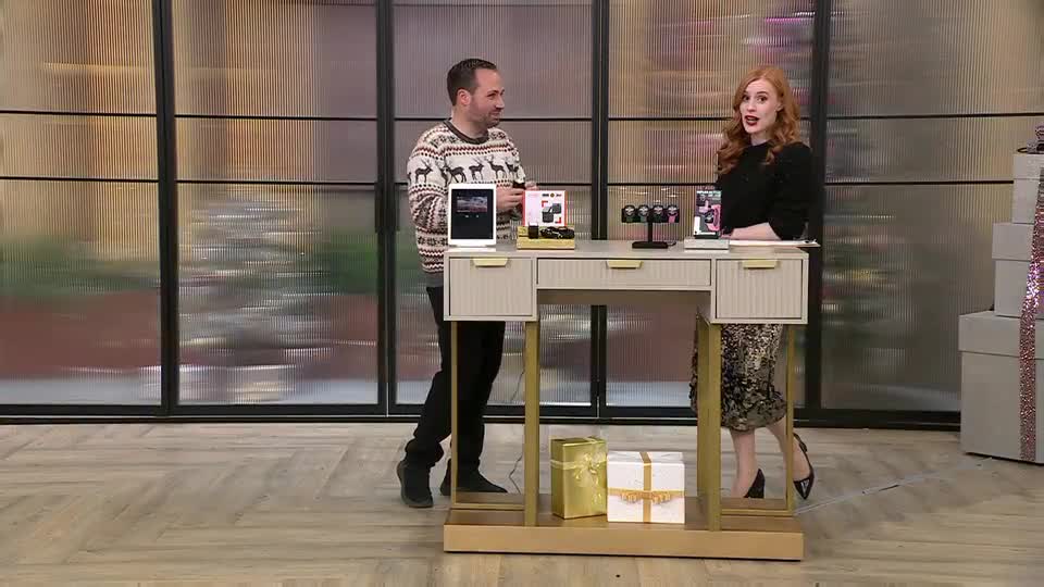 The man in the reindeer sweater gestures towards a tablet on the desk, while the woman in the black top and patterned skirt stands beside him, speaking. They're on a QVC Extra set in the UK, surrounded by gift boxes.
