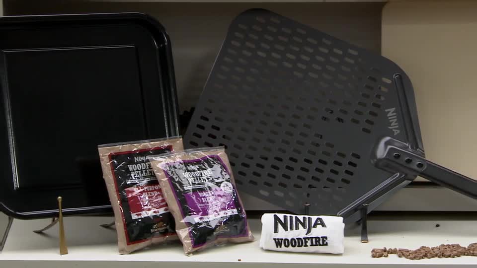 A black pizza oven tray and a perforated pizza peel stand beside two bags of wood pellets. Beside them is a small towel with "NINJA WOODFIRE" printed on it.

