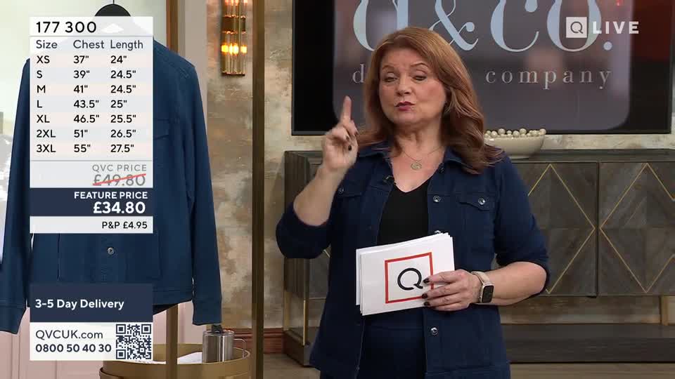 A woman on QVC Extra holds up a stack of cards, one with a prominent red "Q" on it, while gesturing with her other hand. Beside her, a dark blue jacket hangs, displaying a size chart and pricing information.
