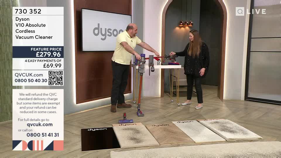 A man in a yellow shirt demonstrates a Dyson vacuum cleaner, while a woman in a black sequined top stands beside him. Five different carpet swatches, each showing varying amounts of debris, lie on the floor in front of them.
