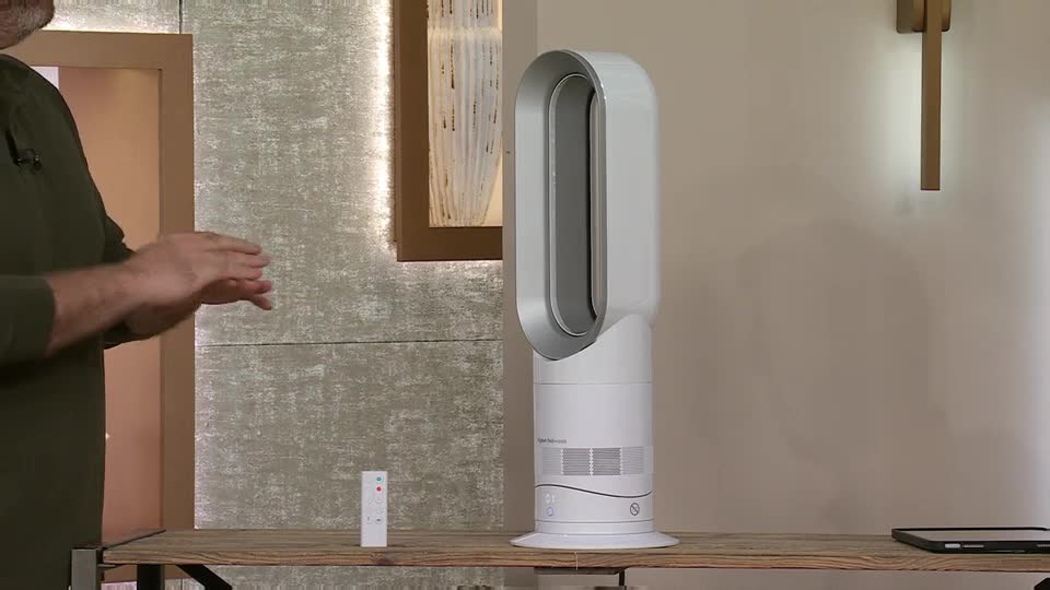 A man in a green shirt gestures with his hands, a small white remote control resting on the table before him. Beside it, a tall white fan stands ready, its sleek design catching the light.