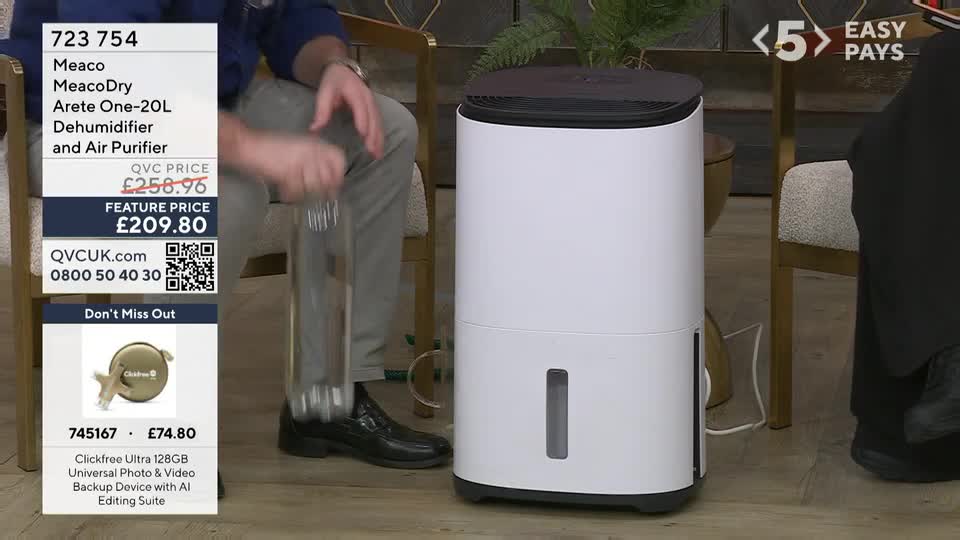 A man in grey trousers and a blue shirt is holding a clear bottle over a tall white dehumidifier. The QVC Extra graphic displays a feature price of £209.80 for the Meaco Dehumidifier and Air Purifier.