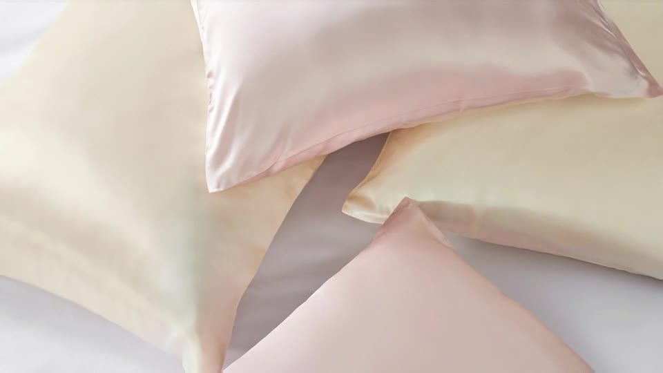 Soft, pale pink and cream pillowcases are layered artfully. The satin fabric catches the light, creating gentle highlights across the smooth surfaces.
