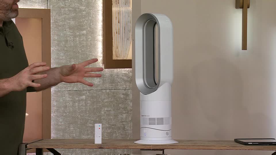 A man gestures with his hands as he talks about the white Dyson fan on the table. A remote control sits on the wooden surface next to the fan.