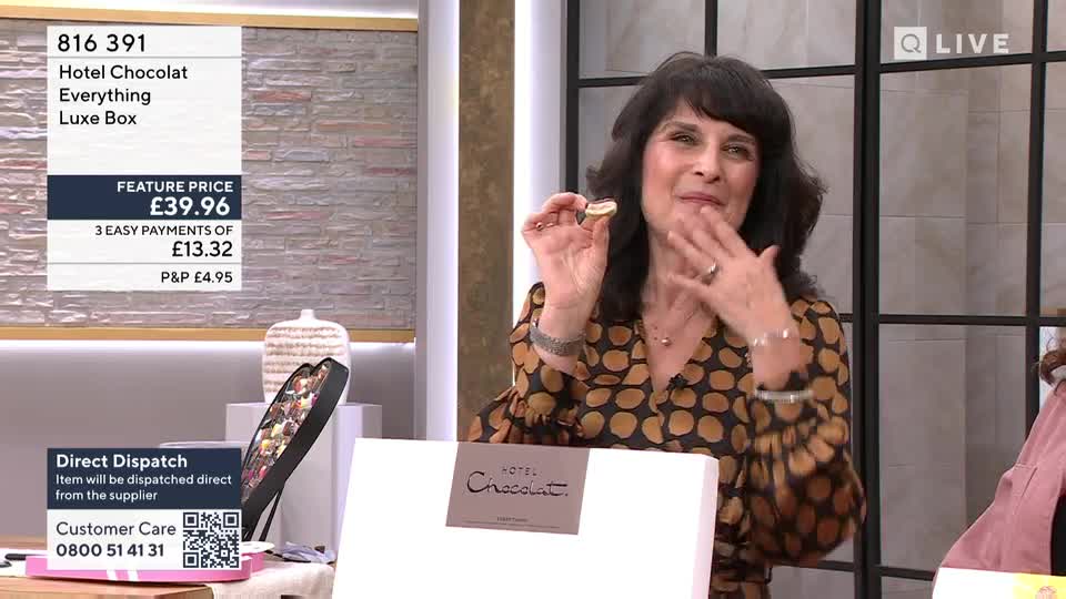 A woman holds up a small, golden chocolate, her fingers touching her lips. Beside her, a large white box with "Hotel Chocolat" printed on it sits open, revealing a selection of chocolates.