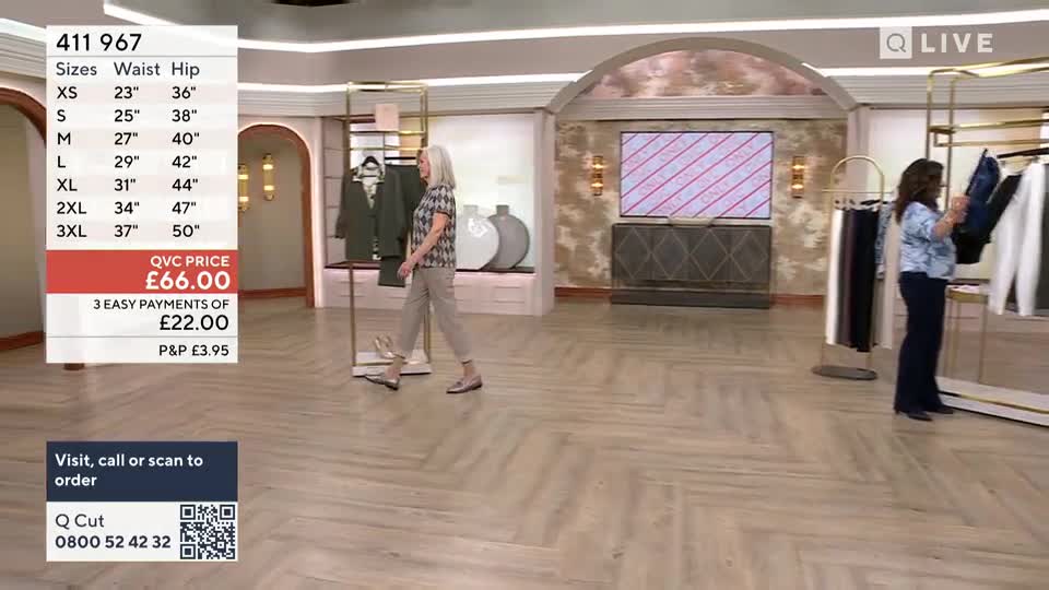 A woman in a patterned shirt and grey trousers walks past a rack of green jackets. Another woman stands by a rack of clothes, examining a dark blue garment.