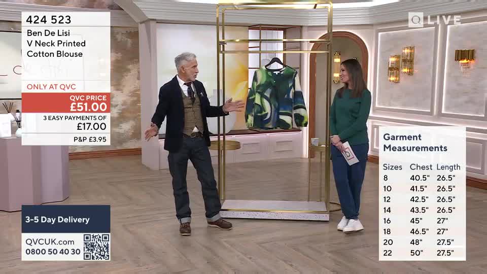 A man with grey hair gestures enthusiastically while a woman stands beside him, holding a card. A vibrant green and blue printed blouse hangs on a display rack between them. A man with grey hair gestures enthusiastically while a woman stands beside him, holding a card. A vibrant green and blue printed blouse hangs on a display rack between them.