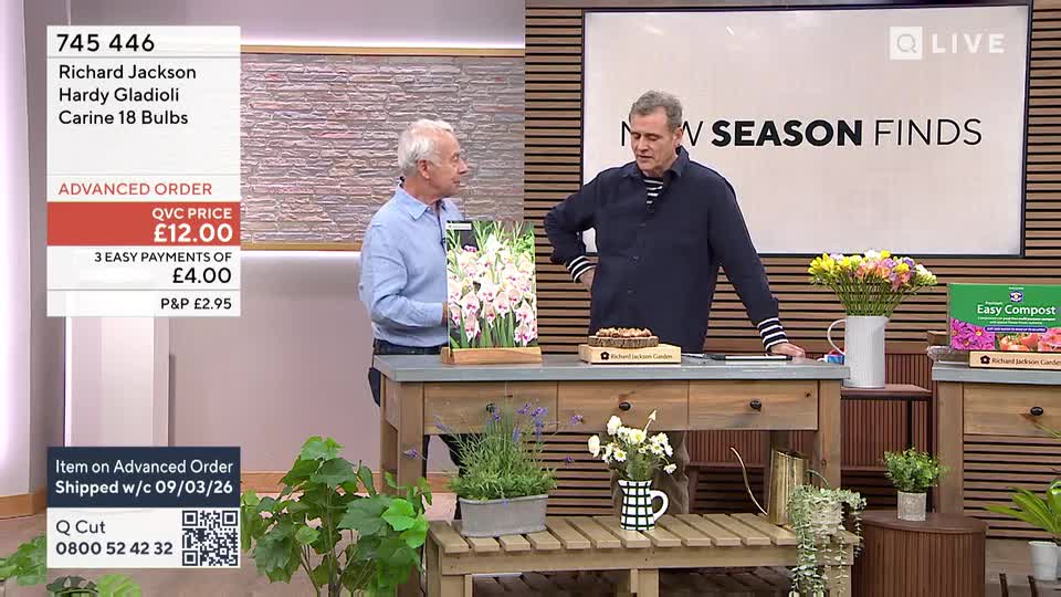 Two men stand on a set, one holding a display of gladioli bulbs. The other gestures towards a box of compost and a vase of flowers on a wooden table. Two men stand on a set, one holding a display of gladioli bulbs. The other gestures towards a box of compost and a vase of flowers on a wooden table.