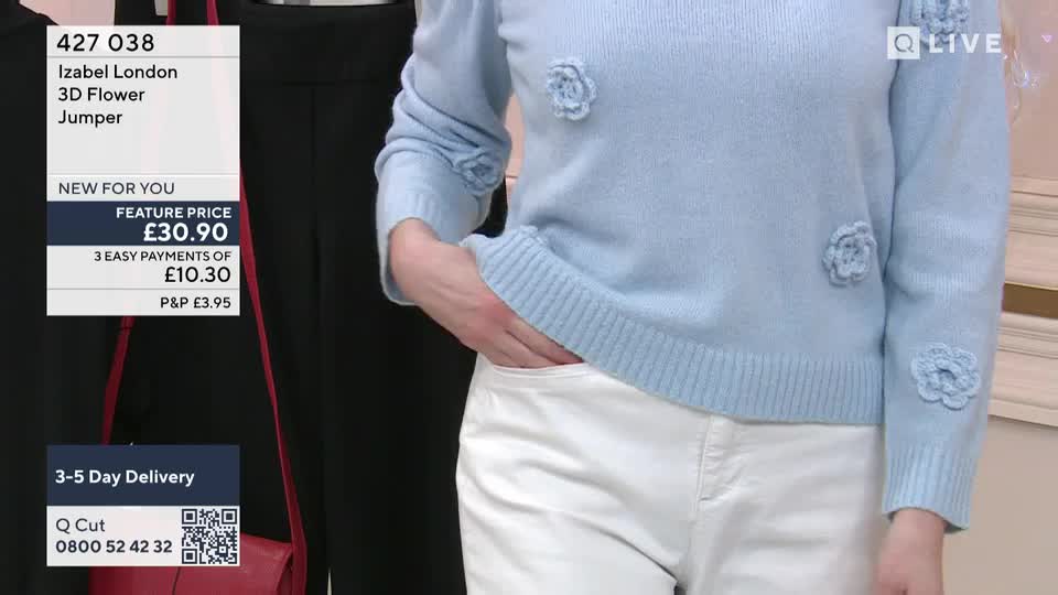 A hand slips into the pocket of white trousers, resting against the pale blue jumper adorned with fabric flowers. The QVC Extra screen displays the item's price and delivery information. A hand slips into the pocket of white trousers, resting against the pale blue jumper adorned with fabric flowers. The QVC Extra screen displays the item's price and delivery information.