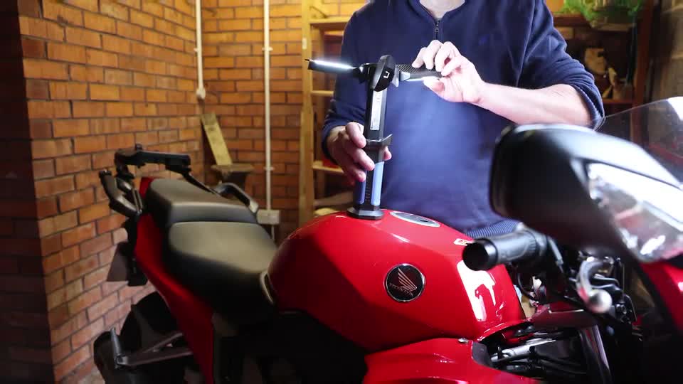 A person is adjusting a bright LED light attached to the handlebars of a red motorcycle. The light beam shines forward, illuminating the bike's fuel tank.