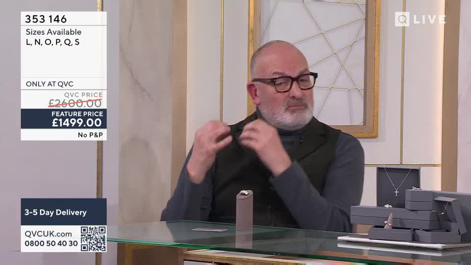 A man with glasses gestures animatedly as he talks about a piece of jewelry. On the table before him, several grey boxes display rings and a delicate necklace.