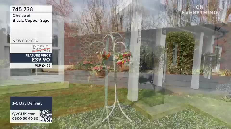 A white metal plant stand with three hanging baskets overflows with colourful flowers. The stand is positioned on a gravel path in front of a brick wall and a small garden building.
