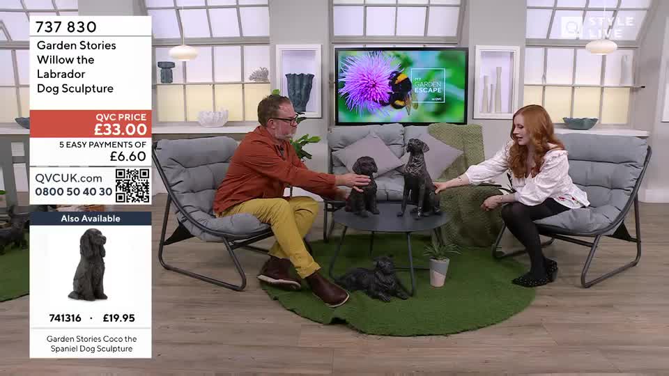 A man in a rust-coloured shirt and yellow trousers gestures towards a small, dark dog sculpture on a coffee table. Beside him, a woman in a white top and black tights reaches out to touch a larger dog sculpture.