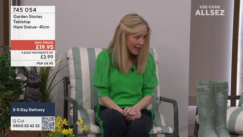 A woman in a bright green jumper is sitting on a striped chair, talking. To her left, a dark grey hare statue stands amongst some greenery.