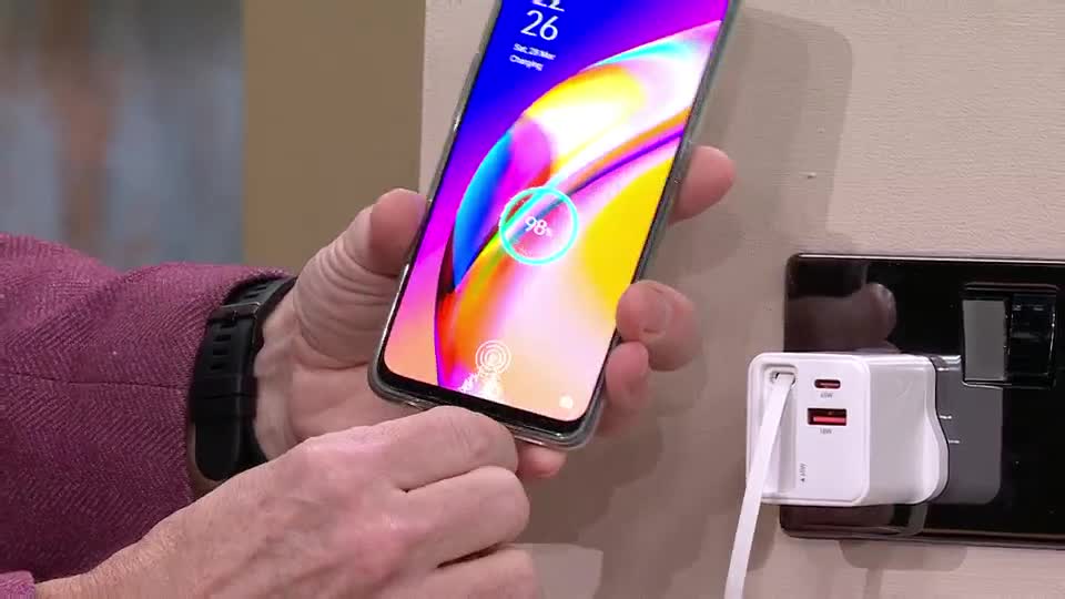 A hand holds a smartphone, its screen displaying a charging icon at 98%. A white charging adapter is plugged into a UK wall socket.