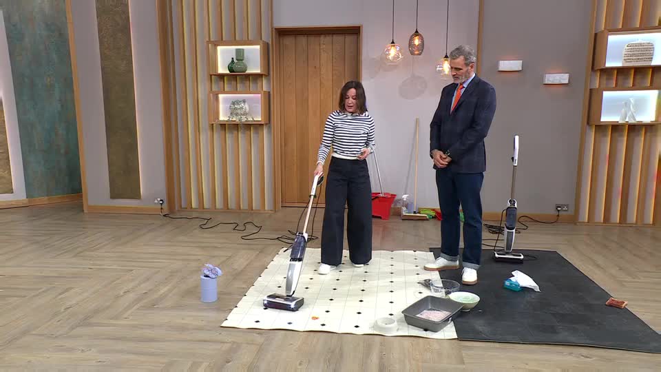 A woman in a striped top and wide-leg trousers pushes a cordless vacuum cleaner across a patterned mat. Beside her, a man in a suit and tie watches, a second vacuum standing nearby.