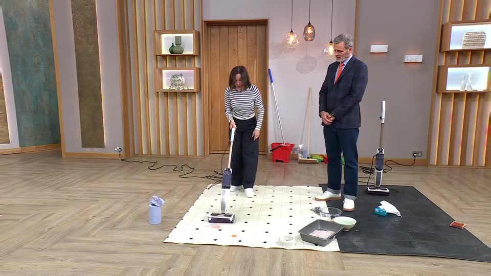 A woman in a striped shirt and dark trousers operates a floor cleaner on a patterned rug. A man in a suit stands nearby, watching the demonstration.