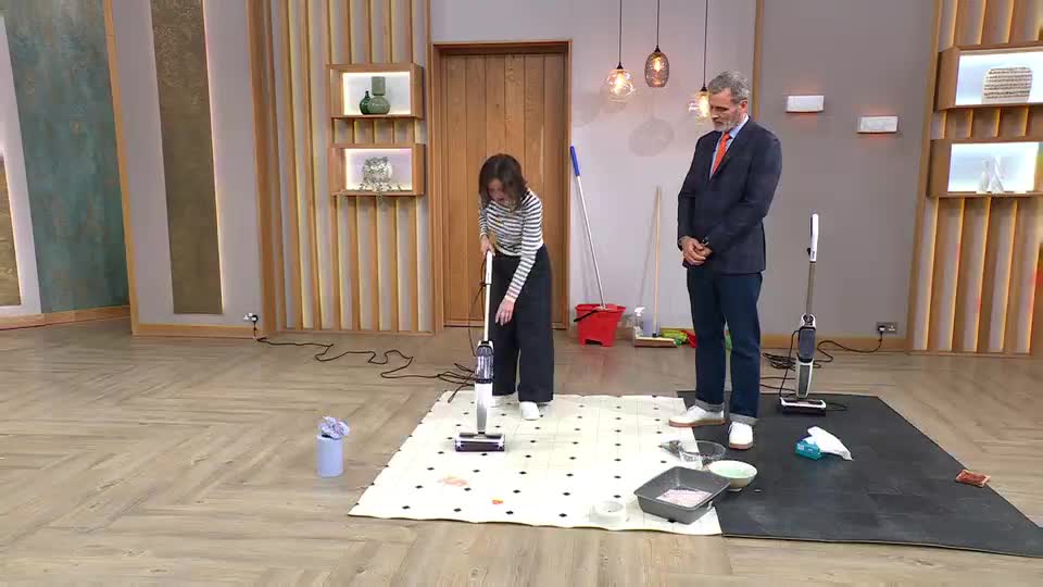 A woman in a striped shirt vacuums a patterned rug, while a man in a suit watches. The demonstration is happening on a studio floor, likely for QVC Extra.