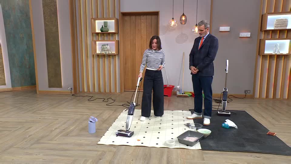 A woman demonstrates a vacuum cleaner on a patterned mat while a man in a suit watches. The vacuum's brush head is actively spinning as she moves it across the floor.
