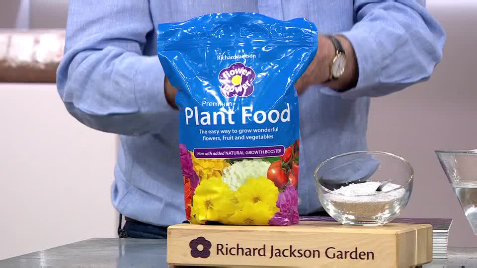 A man in a blue shirt holds a bag of "Flower Power Plant Food." Beside it, a small glass bowl contains a white granular substance.