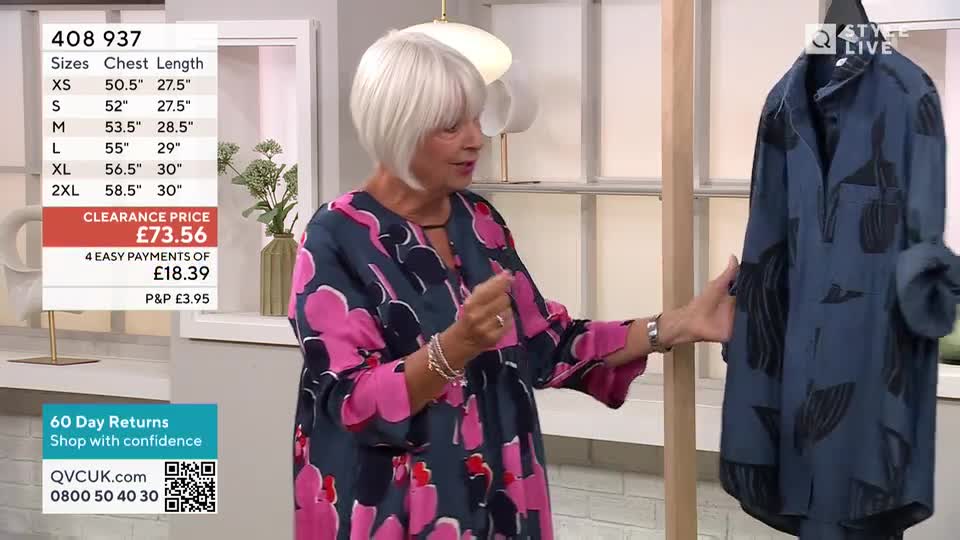 A woman in a patterned dress gestures towards a blue shirt hanging on a rack. She's presenting a garment on a QVC UK show, discussing its features.
