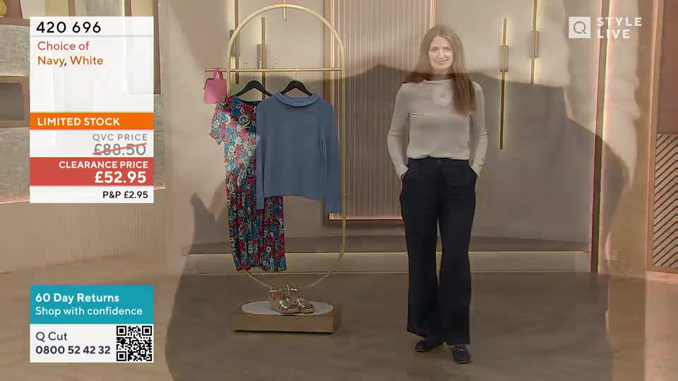 A woman in a light-colored top and dark trousers stands in front of a clothing rack, smiling at the camera. The screen displays a "Limited Stock" sale for a navy and white item, with a QVC price and a clearance price.
