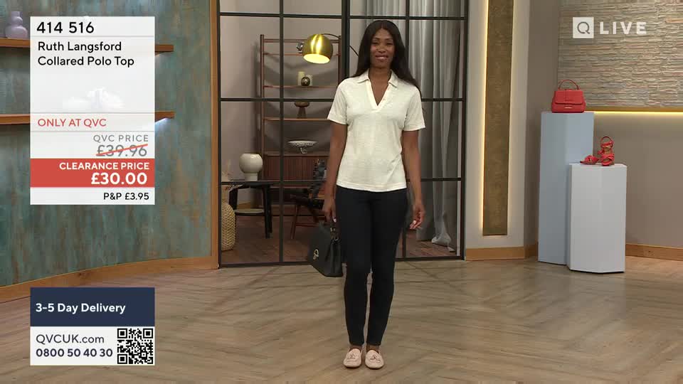 A woman in a collared polo top and black trousers stands center stage, smiling directly at the camera. Behind her, a display shows a handbag and shoes, all part of the QVC presentation.

