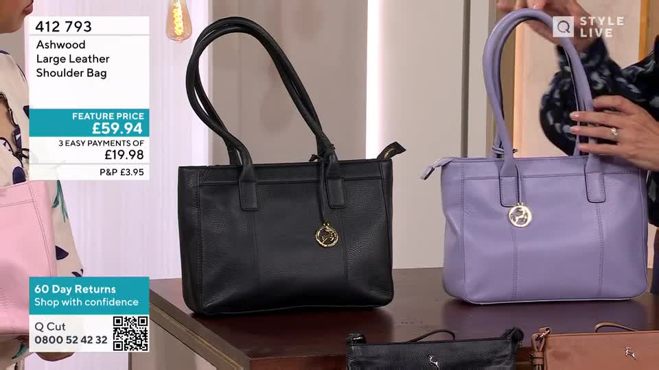 A woman holds up a lavender leather shoulder bag, showing off its shape and detailing. Beside it, a black bag with long handles sits on a table, with the price displayed on the screen.
