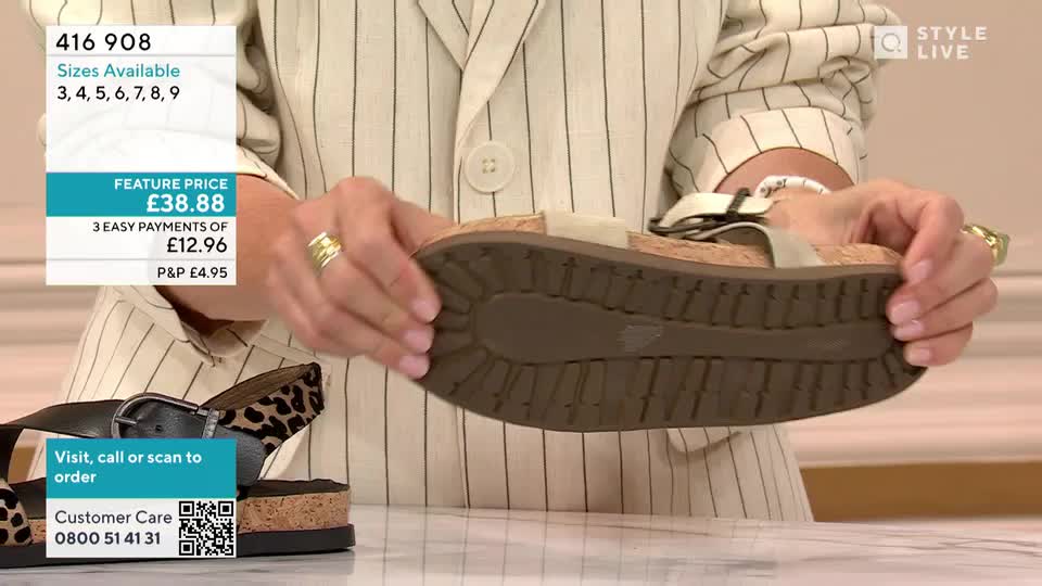 A presenter in a pinstripe jacket holds up a sandal, showing the textured sole. The camera focuses on the shoe's underside as she demonstrates its features for a QVC-style broadcast.
