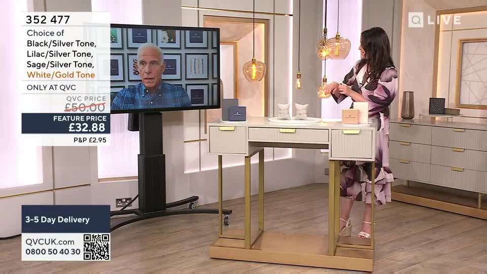 A woman in a floral dress, standing at a display table, adjusts a bracelet. On a screen behind her, a man in a blue shirt is talking, while the QVC price and details are displayed on the left.
