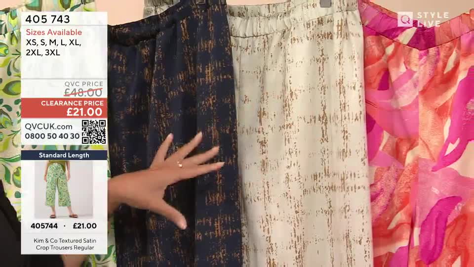 A presenter is holding up a selection of patterned trousers, showing them off for a QVC Style segment. The dark blue pair has a gold-toned grid pattern, and the off-white pair has a similar design.
