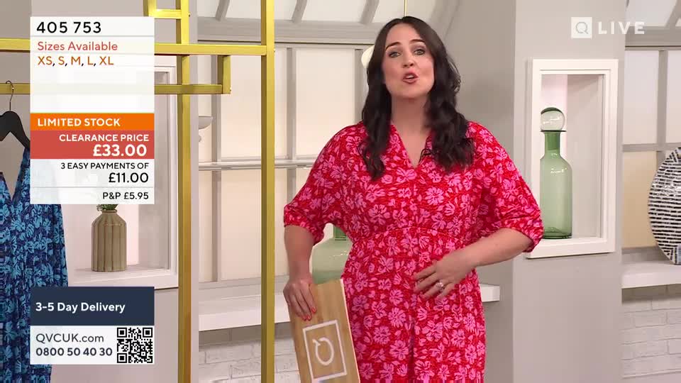 A woman in a bright floral dress stands in a studio, gesturing towards her stomach while talking. A screen displays details about the dress, including the clearance price in pounds.
