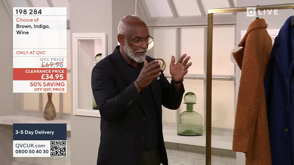 The man in the black jacket gestures with his hands, discussing a product on a QVC-style show. Behind him, a rack displays coats in various colors, and a graphic advertises a clearance price.
