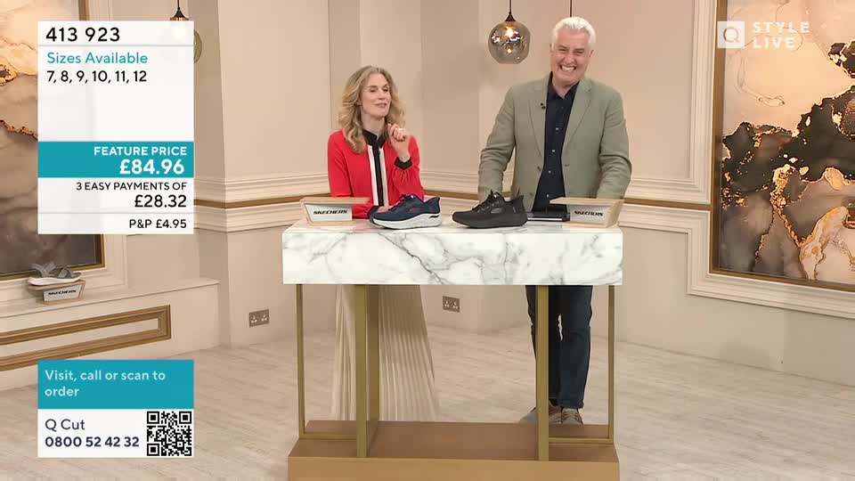A woman in a red jacket gestures towards a pair of blue trainers on a marble-topped table. Her co-host, grinning, stands next to a black pair of trainers, ready to talk about the footwear.
