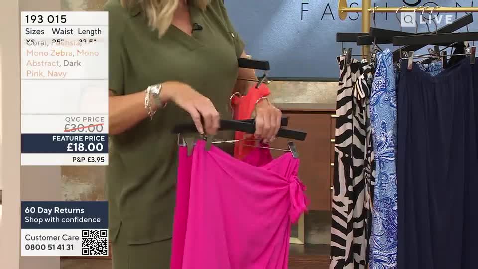 A presenter in a green top holds up a vibrant pink skirt, showing it off to the camera. Behind her, other skirts hang on a rack, ready to be featured on the QVC show.
