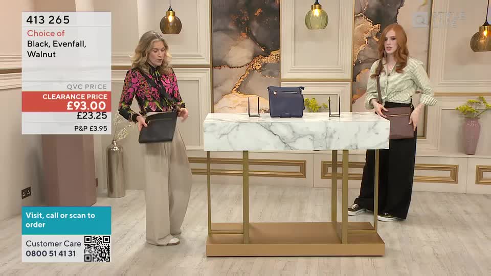 Two women stand in a studio, each holding a crossbody bag, demonstrating the different styles and colours available. The host on the left gestures to her black bag, while the other model displays a brown one, both standing near a display table.
