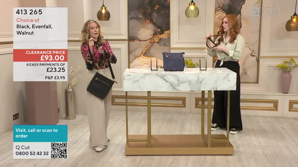 A woman in a floral top holds a black bag, showing it off as she stands near a marble-topped table. Across from her, another woman holds a bag's strap, likely demonstrating its features for a QVC-style broadcast.
