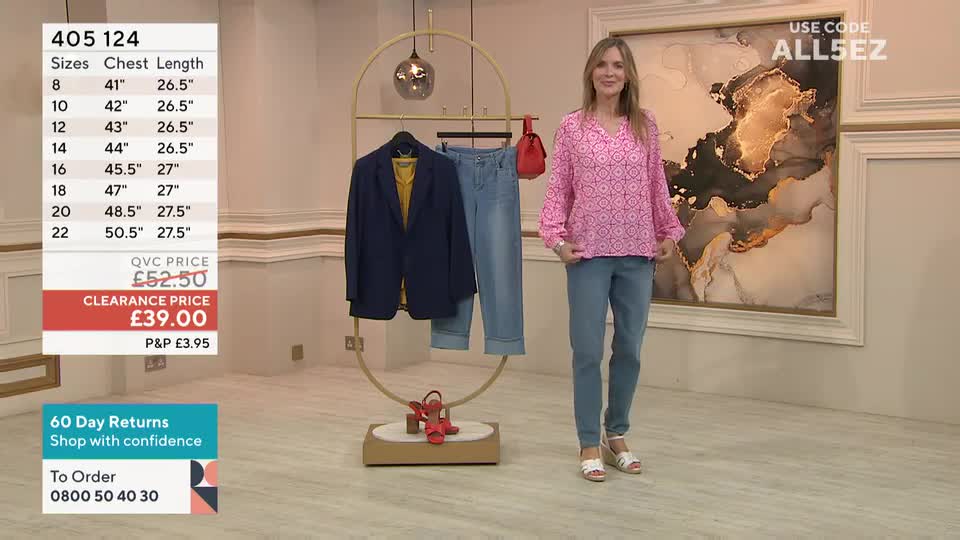 A woman in a pink patterned top and jeans stands in front of a display of clothes, smiling at the camera. Behind her, a navy blazer and jeans hang on a golden rack, and a price tag flashes on the screen.
