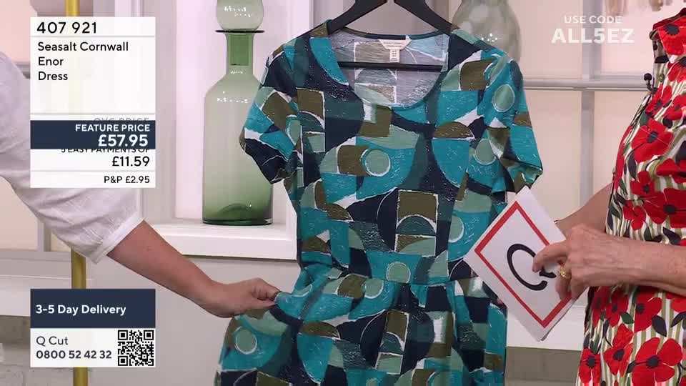 A patterned dress hangs on a rack, its fabric being gently pulled out by a presenter. Another woman holds up a card with a "C" on it, while a price tag and promotional information are displayed on the screen.

