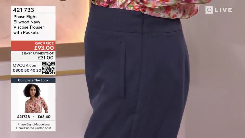 The presenter is showing off the navy trousers, pointing out the pocket detail. The camera is zoomed in on the trousers as the price flashes on the screen in the background.
