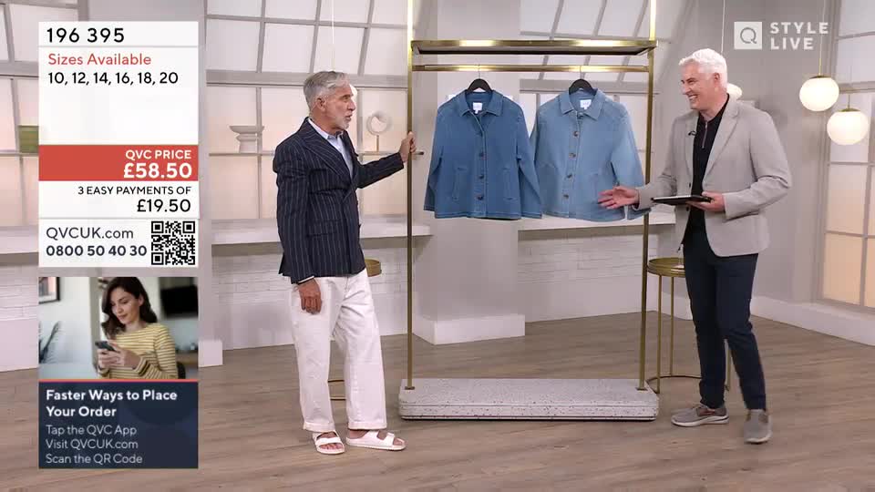 Two men stand in a studio, one in a pinstripe blazer gesturing towards a rack of denim jackets. The other man, holding a tablet, is smiling and pointing towards the jackets as well.
