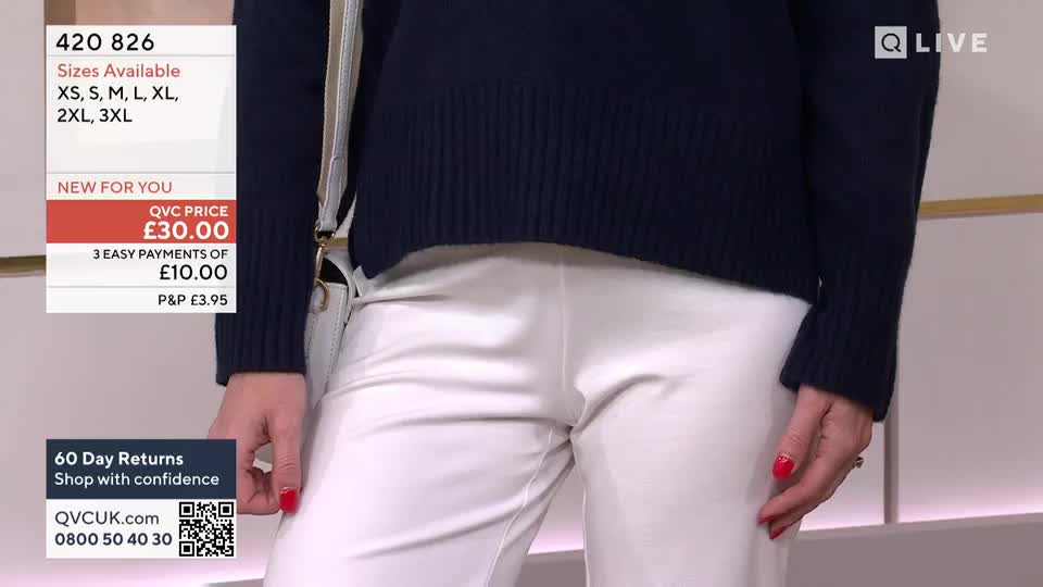 A woman stands, hands resting at her sides, showcasing a navy sweater and white trousers. The QVC price of £30.00 is displayed, and the presenter's nails are painted a bright red.
